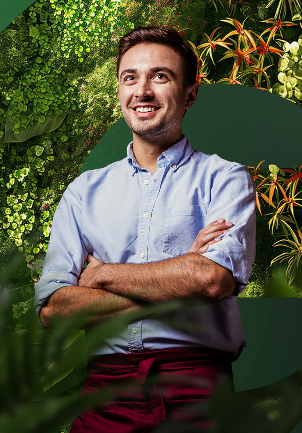 Rapaz de cabelo castanho, com uma camisa azul e calças bordeaux, de braços cruzados à frente de um fundo de cor verde e conjuntos de plantas