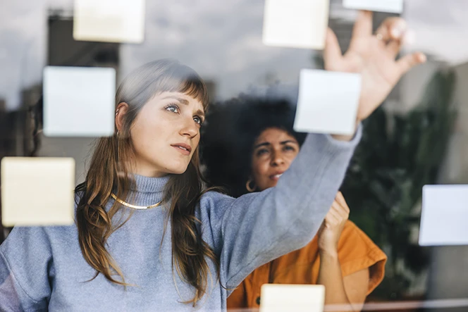 2 mulheres reunidas em frente a quadro, com post-its