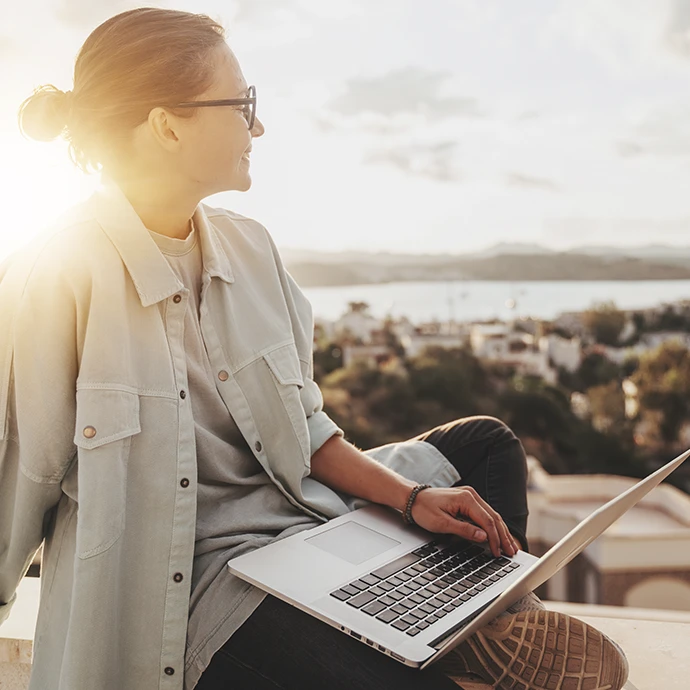 Mulher sentada, ao pôr do sol em miradouro e a sorrir, com o computador no colo