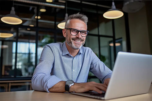 smiling man with a beard and glasses, sitting at a desk with an open laptop