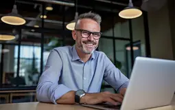 homem com barba e óculos, sorridente, sentado numa secretária, com portátil aberto
