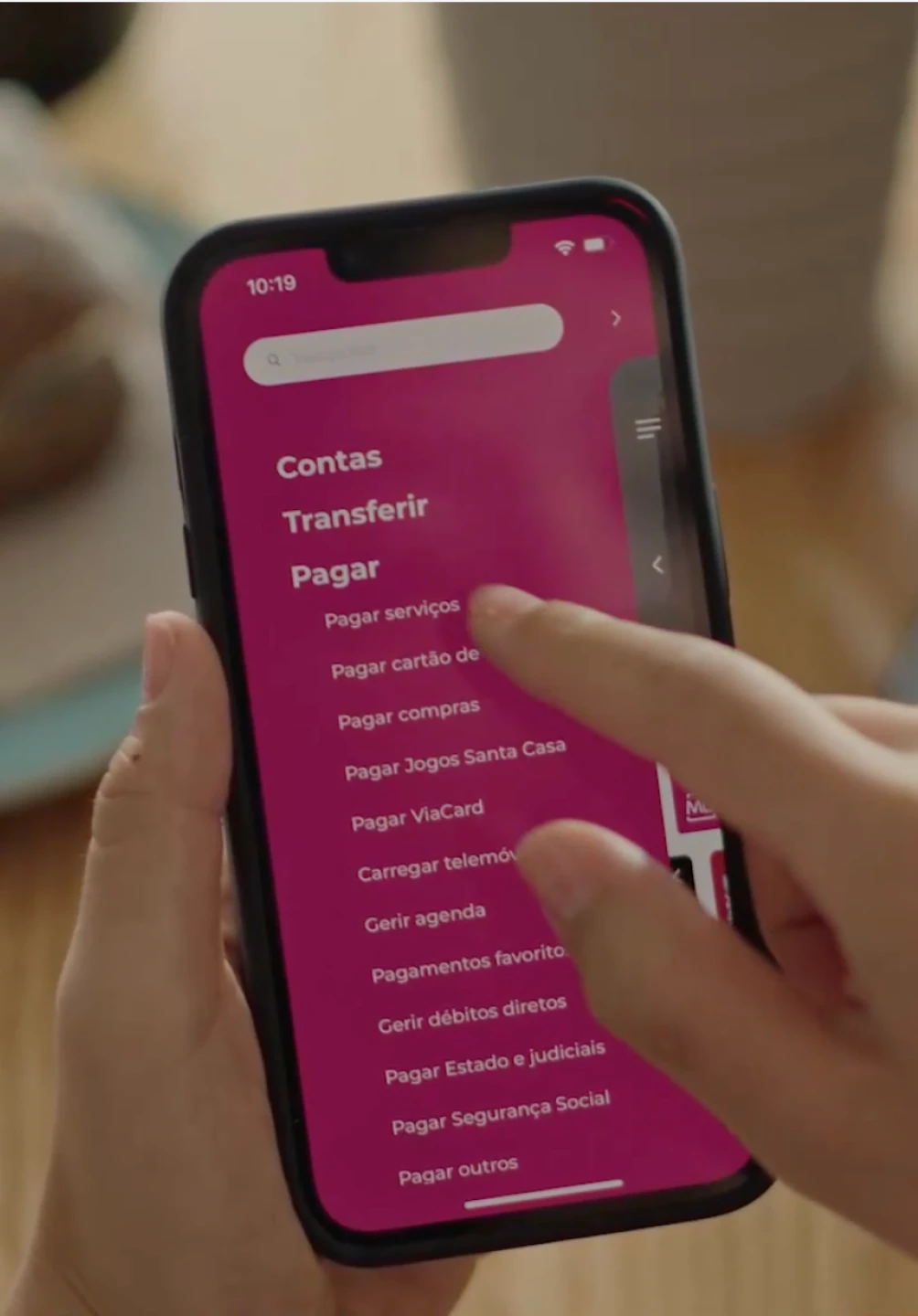 Mão a agarrar um telemóvel que no seu ecrã apresenta o menu da App Millennium, com destaque para a secção "Pagar"