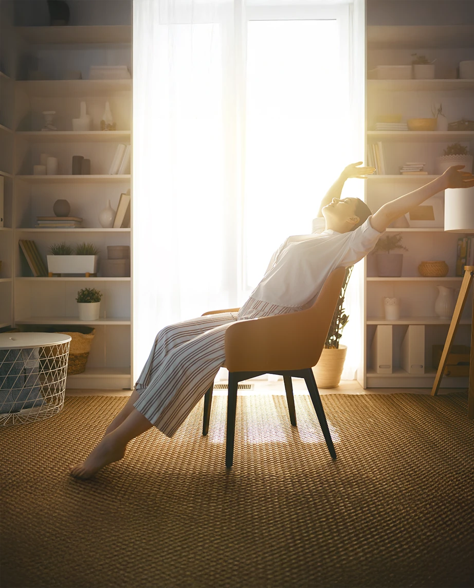 mulher sentada numa poltrona na sala, a espreguiçar-se, com luz do sol a entrar pela grande janela