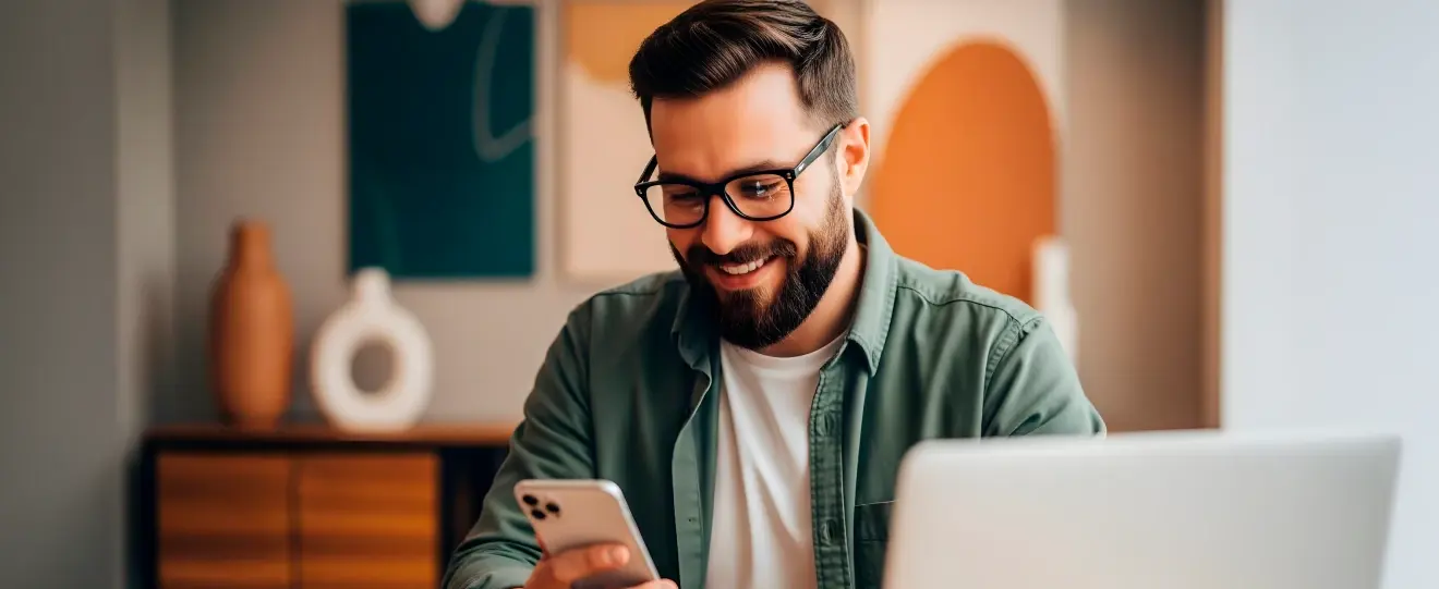 Homem de óculos sorridente a olhar para o telemóvel, com portátil