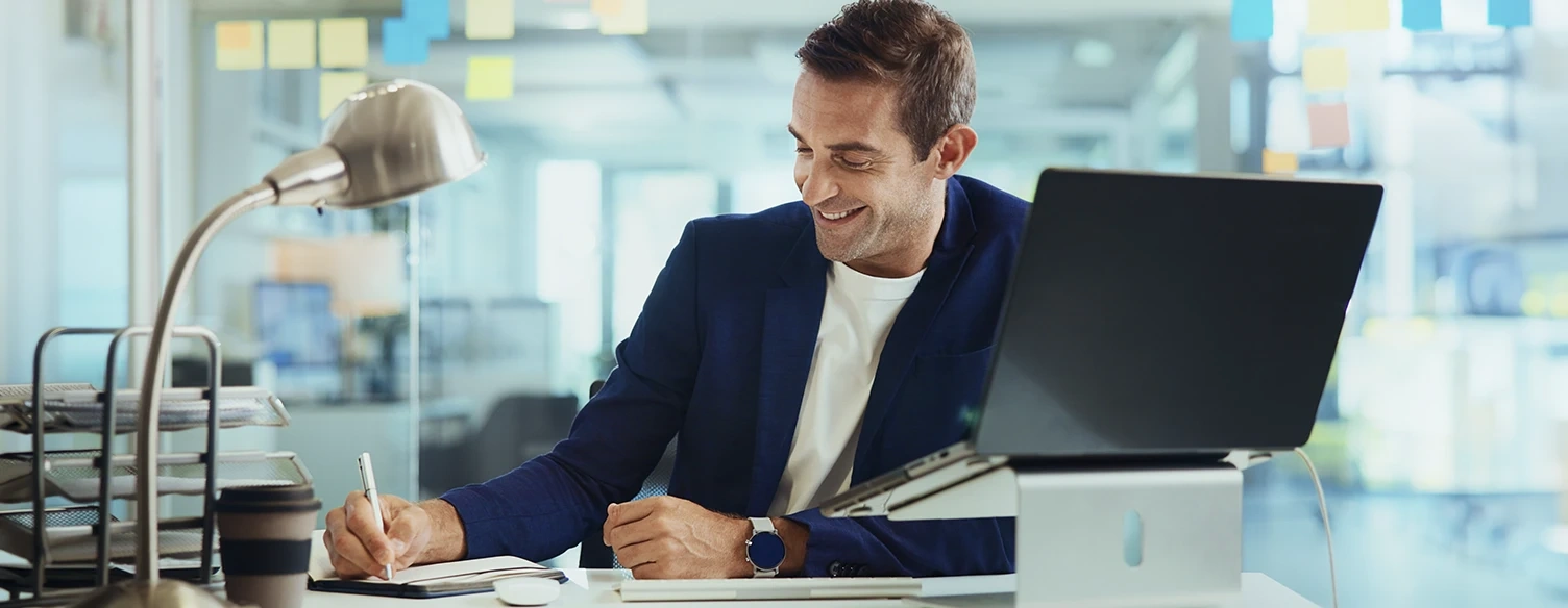 Homem sorridente a escrever num caderno numa secretária de escritório