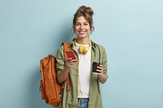 rapariga sorridente, com mochila, telemóvel na mão, e headphones à volta do pescoço