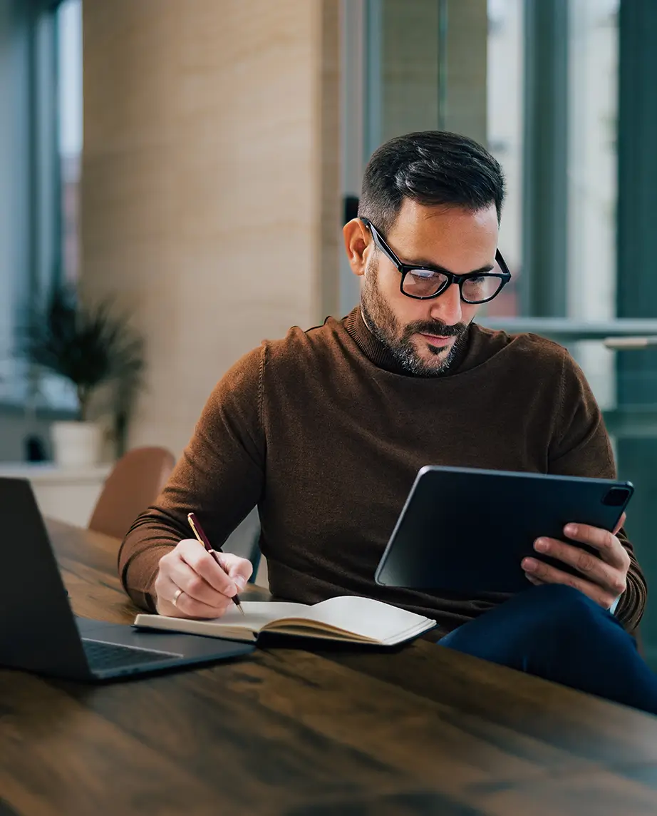 man sitting at the desk, writing notes and consulting a tablet