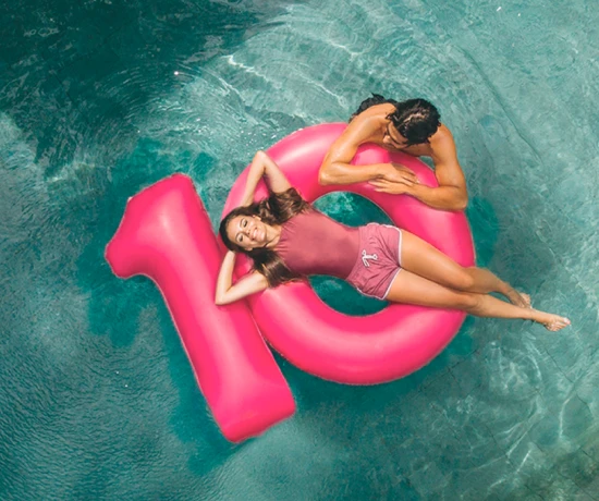 Duas pessoas numa piscina com boias cor-de-rosa em forma de 10, carrinho de compras e posto de abastecimento de combustível