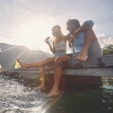 Casal sentado na borda de um barco com os pés na água ao pôr do sol