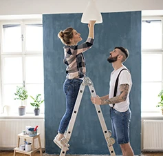 couple, she on a stepladder changing a light bulb in a lamp, he holding the stepladder