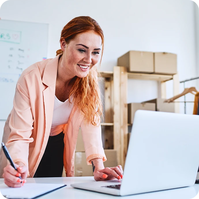 senhora sorridente, com computador e caneta na mão, a escrever num caderno