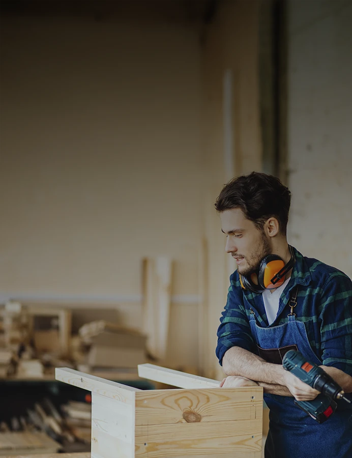 Jovem rapaz serralheiro a construir uma cadeira de madeira, na oficina, vestido de fato macaco de ganga e com perfuradora de bateria na mão