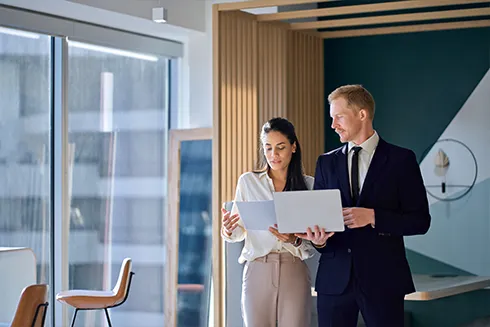 man and a woman in an office, holding a laptop and papers