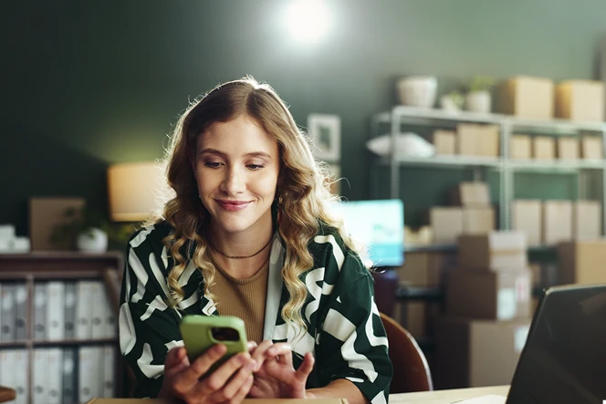 senhora sorridente num escritório, com estantes e caixas, com telemóvel na mão