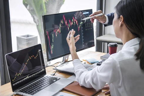 2 pessoas num escritório, com 2 computadores com gráficos, a analisar mercados