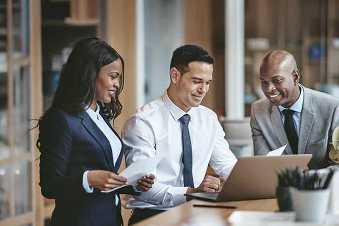 2 men and 1 woman, smiling, in a meeting room with a desk holding a laptop and papers