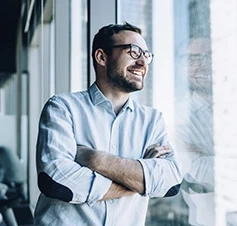 Man with glasses smiling with arms crossed by a window
