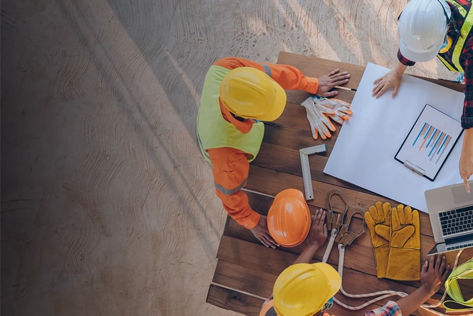 top view of construction workers around a table with a laptop, blueprints, gloves, and helmets