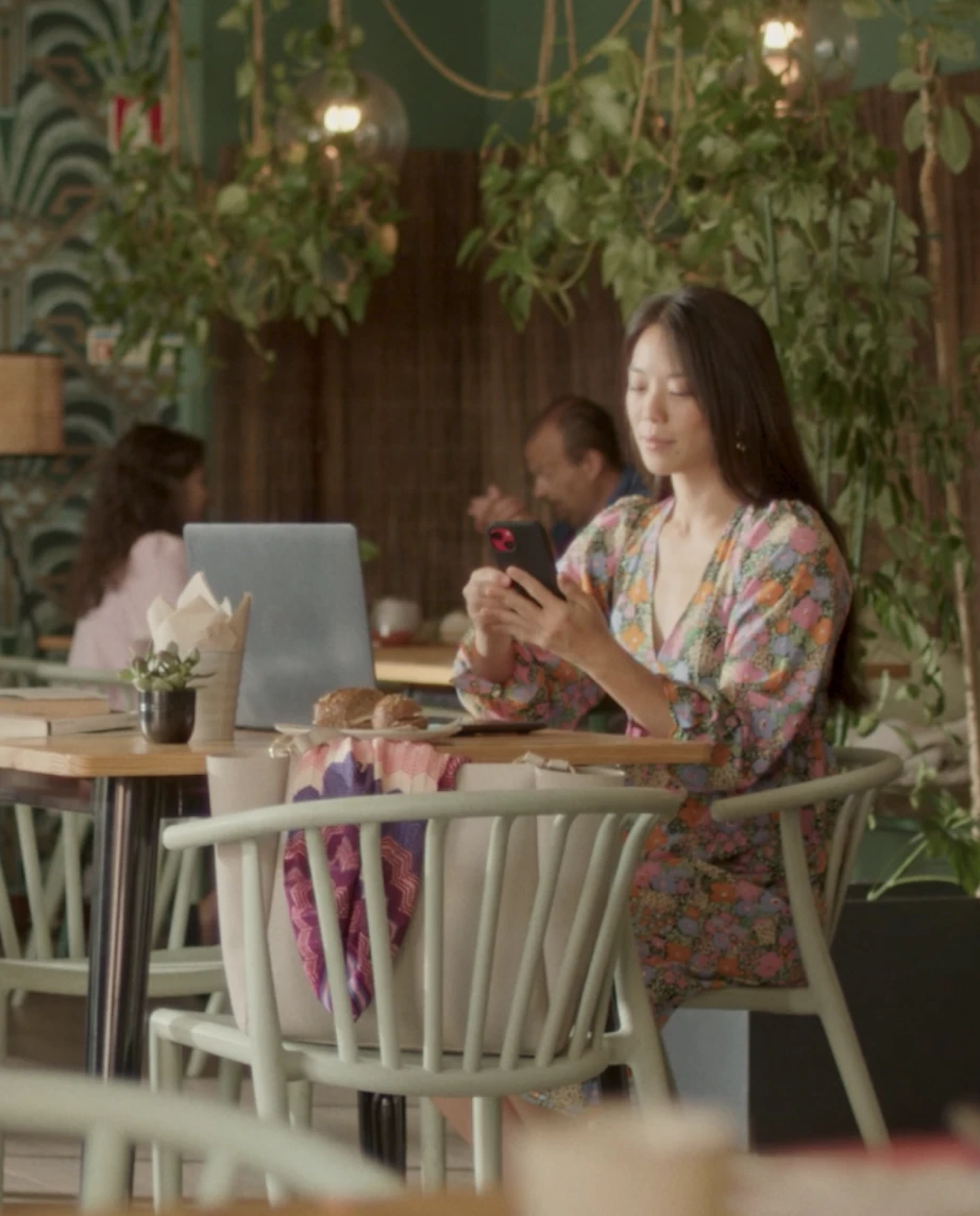 Rapariga na casa dos 30 anos com um vestido com flores a olhar para o seu telemóvel e computador portátil, numa mesa de um café