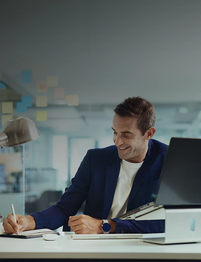 homem sorridente atrás de uma secretária, a escrever num caderno, com computador ao lado