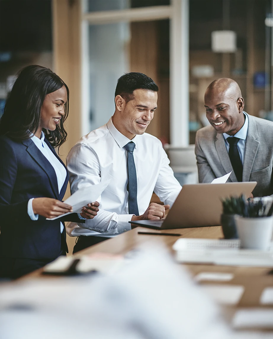 2 homens e 1 mulher, sorridentes, numa sala de reuniões com uma secretária com portátil e papéis