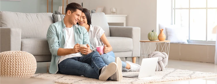 Casal jovem feliz, sentado no chão, segurando canecas e olhando para um portátil