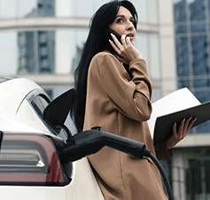 senhora a falar ao telemóvel e com caderno na mão, encostada a carro elétrico em carregamento
