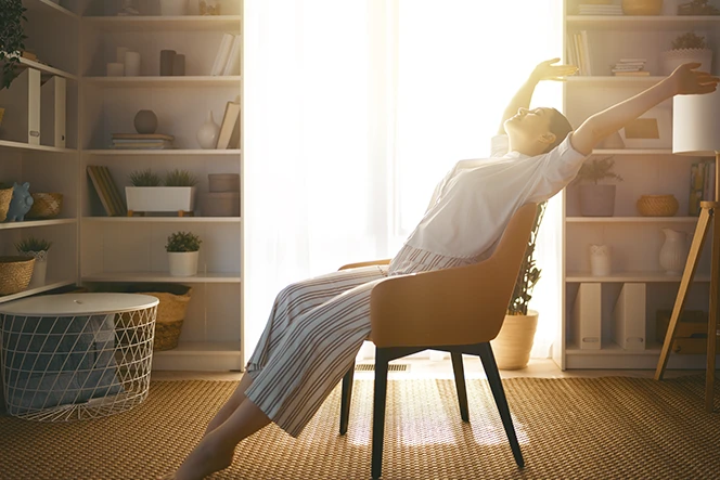 mulher sentada numa poltrona na sala, a espreguiçar-se, com luz do sol a entrar pela grande janela