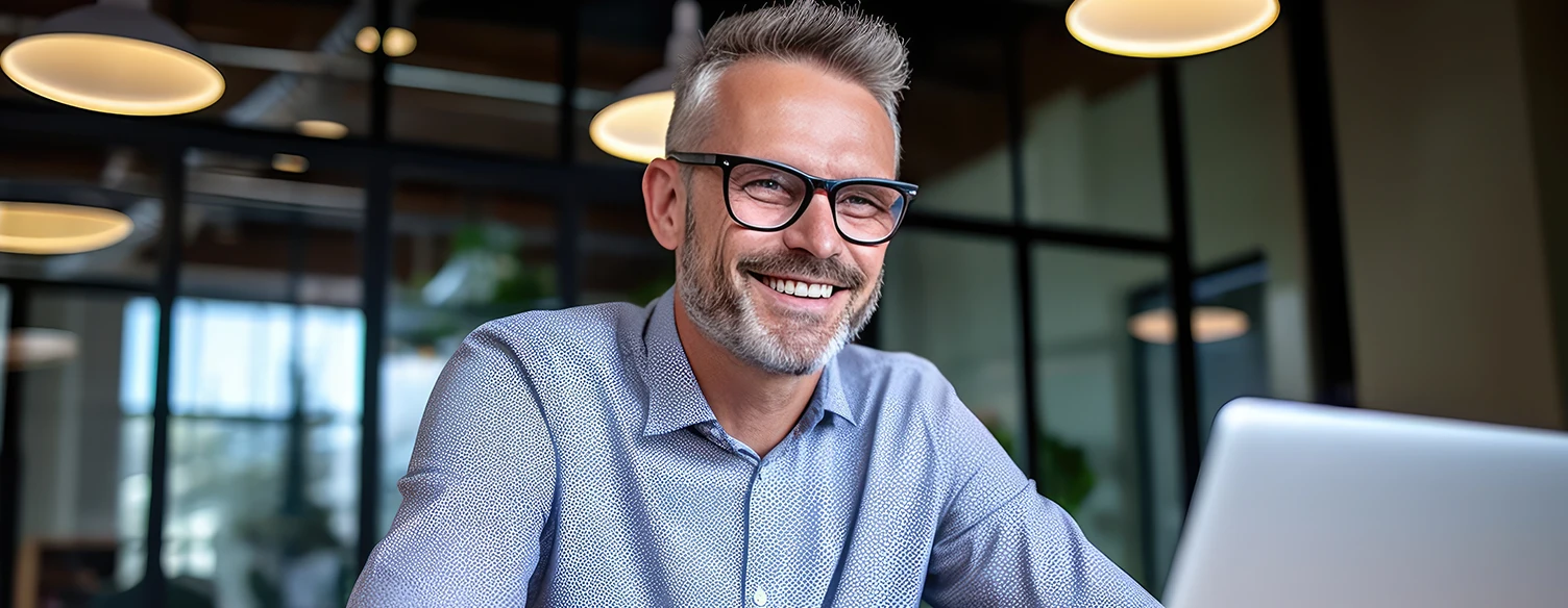 smiling man with a beard and glasses, sitting at a desk with an open laptop