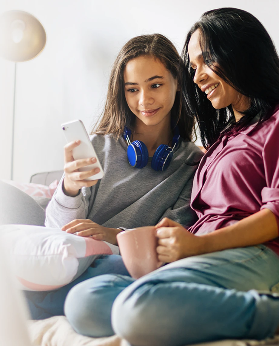 mother holding a cup of tea and the teenage daughter with a phone, sitting on the sofa in a moment of closeness