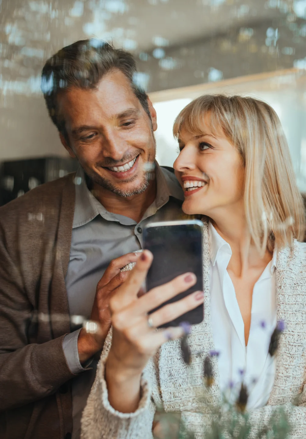 Casal sorridente a olhar para um telemóvel juntos, refletidos numa janela