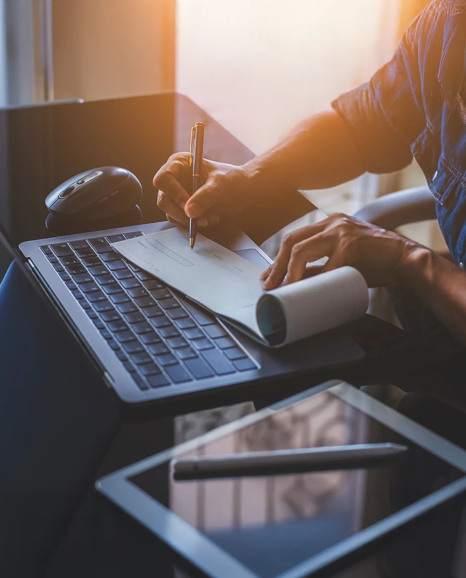 pormenor de mãos a assinar cheque, numa secretária com computador, tablet e caneta, com luz de pôr do sol