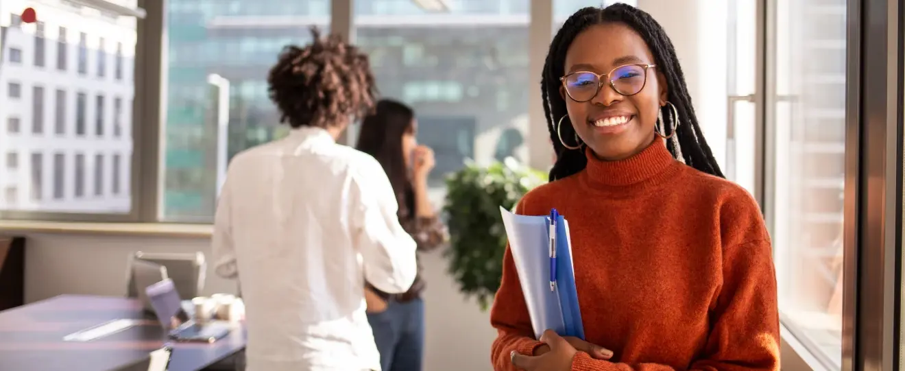 Mulher sorridente com óculos e tranças a segurar uma pasta num escritório