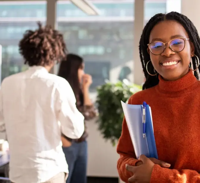 Mulher sorridente com óculos e tranças a segurar uma pasta num escritório