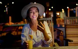 mulher sentada numa mesa de esplanada, à noite, a sorrir, com telemóvel na mão