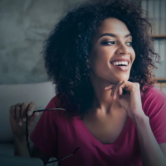 Mulher sorridente de cabelo encaracolado, com blusa rosa, a segurar óculos, a olhar para a direita