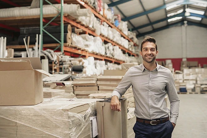 Homem sorridente num armazém, encostado a caixas e prateleiras de produtos