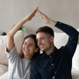 Casal sentado no chão, a sorrir e a fazer forma de casa com os braços