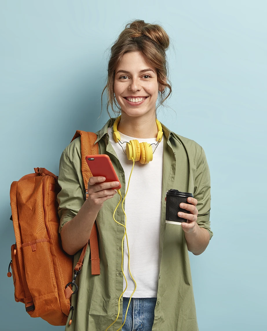 rapariga sorridente, com mochila, telemóvel na mão, e headphones à volta do pescoço