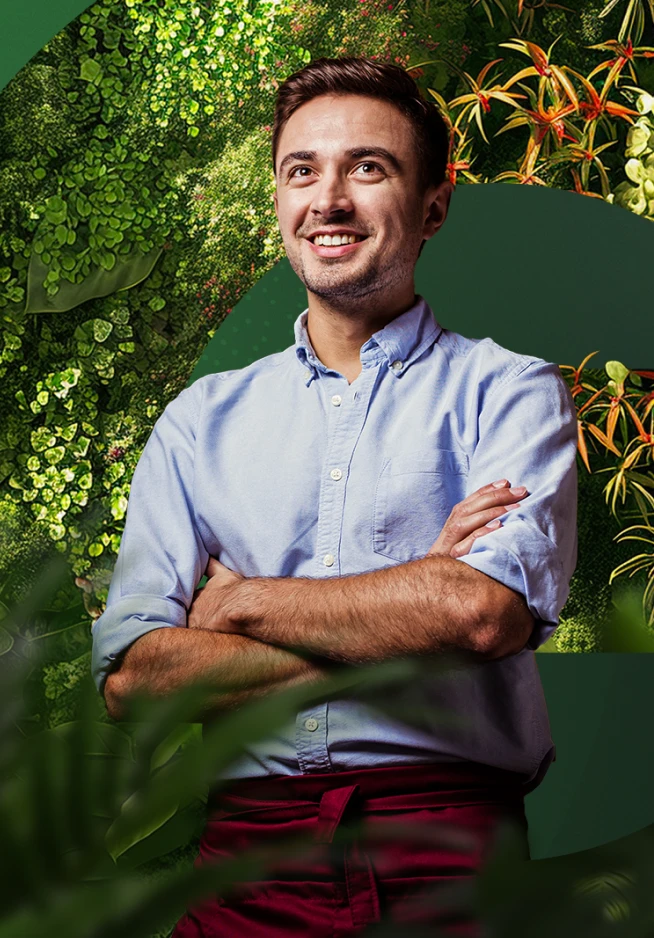 Rapaz de cabelo castanho, com uma camisa azul e calças bordeaux, de braços cruzados à frente de um fundo de cor verde e conjuntos de plantas