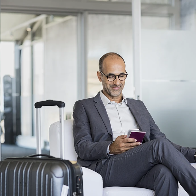Senhor sentado, em sala de espera do Aeroporto, com bagagem e a sorrir com o telemóvel na mão