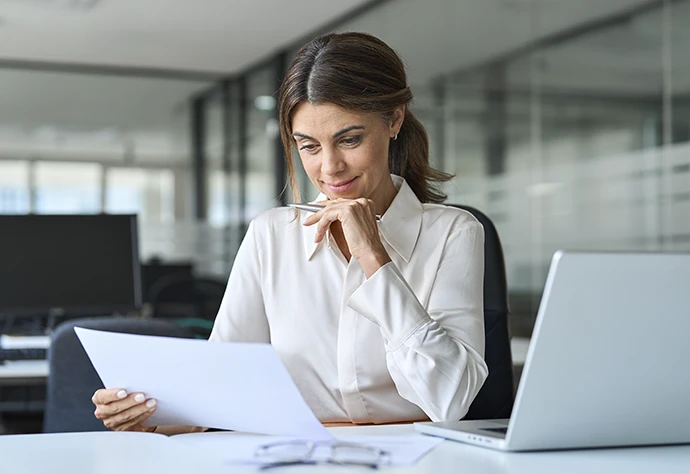 senhora sorridente num escritório, comcomputador ao lado, a ler documento e com caneta na mão