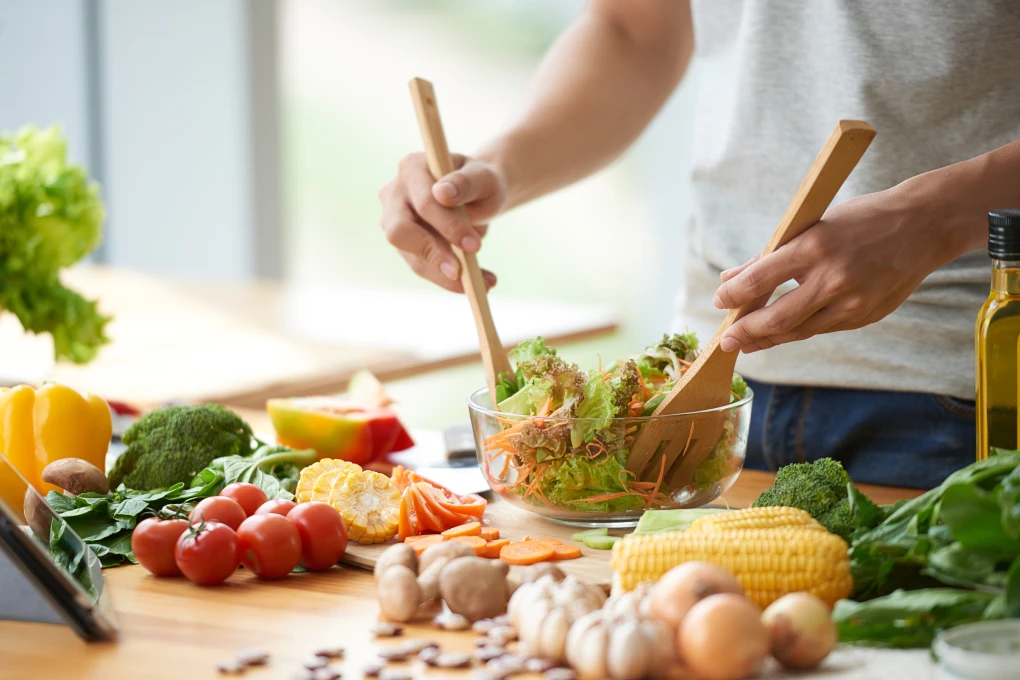 Bancada de cozinha de madeira com vários ingredientes em cima da mesa e uma taça com salada que está a ser mexida