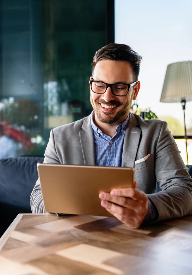 Homem com óculos pretos, camisa azul e branca aos xadrez e blazer cinza, a sorrir para o tablet que tem na mão direita