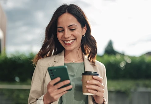 Mulher sorridente a usar o telemóvel enquanto segura num copo de café