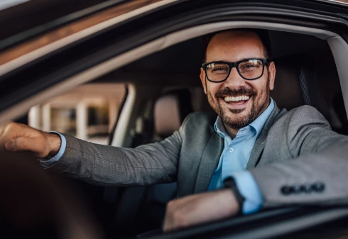 Homem de camisa azul e blazer cinza ao volante de um carro a sorrir pela janela