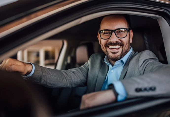 Homem de camisa azul e blazer cinza ao volante de um carro a sorrir pela janela