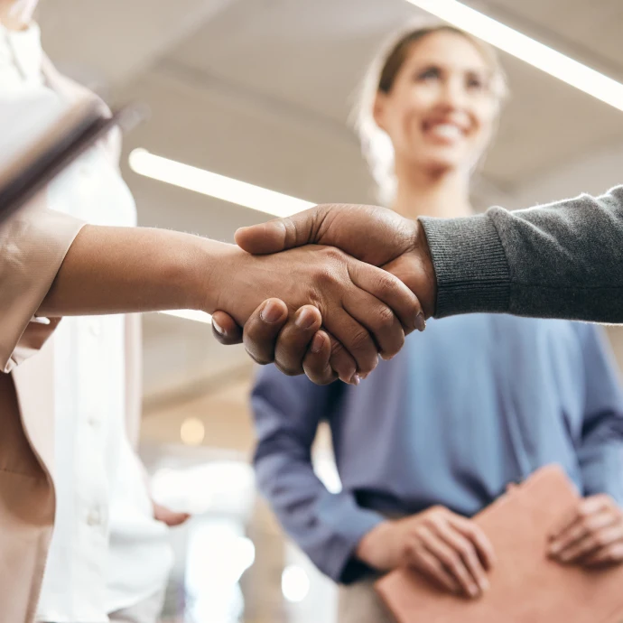Aperto de mãos entre duas pessoas, com uma mulher sorridente em segundo plano