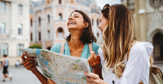 Duas mulheres jovens a sorrir e a segurar um mapa de papel enquanto exploram uma praça europeia com edifícios históricos ao fundo.