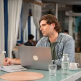 Jovem rapaz, sentado em mesa de esplanada, com o computador aberto e a tirar apontamentos em caderno.